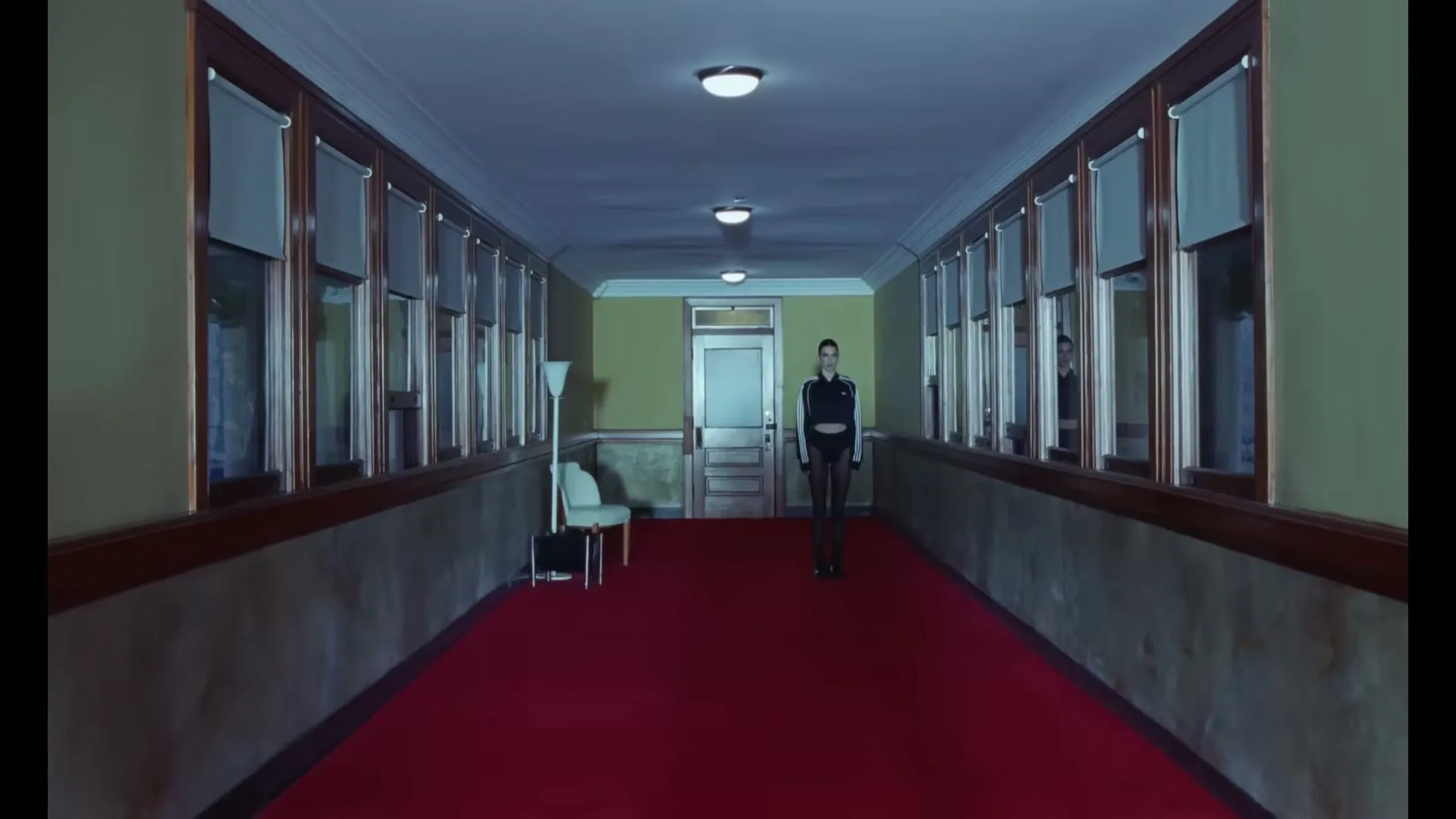 A young woman in a black and white tracksuit stands alone in a long, empty hallway with a red carpet and many windows.