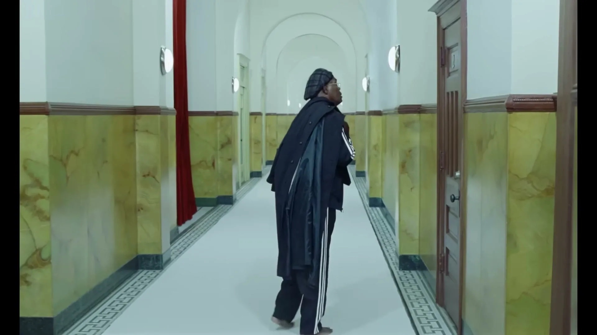 A man in a black coat and track pants walks barefoot down a long, ornate hallway, looking over his shoulder.