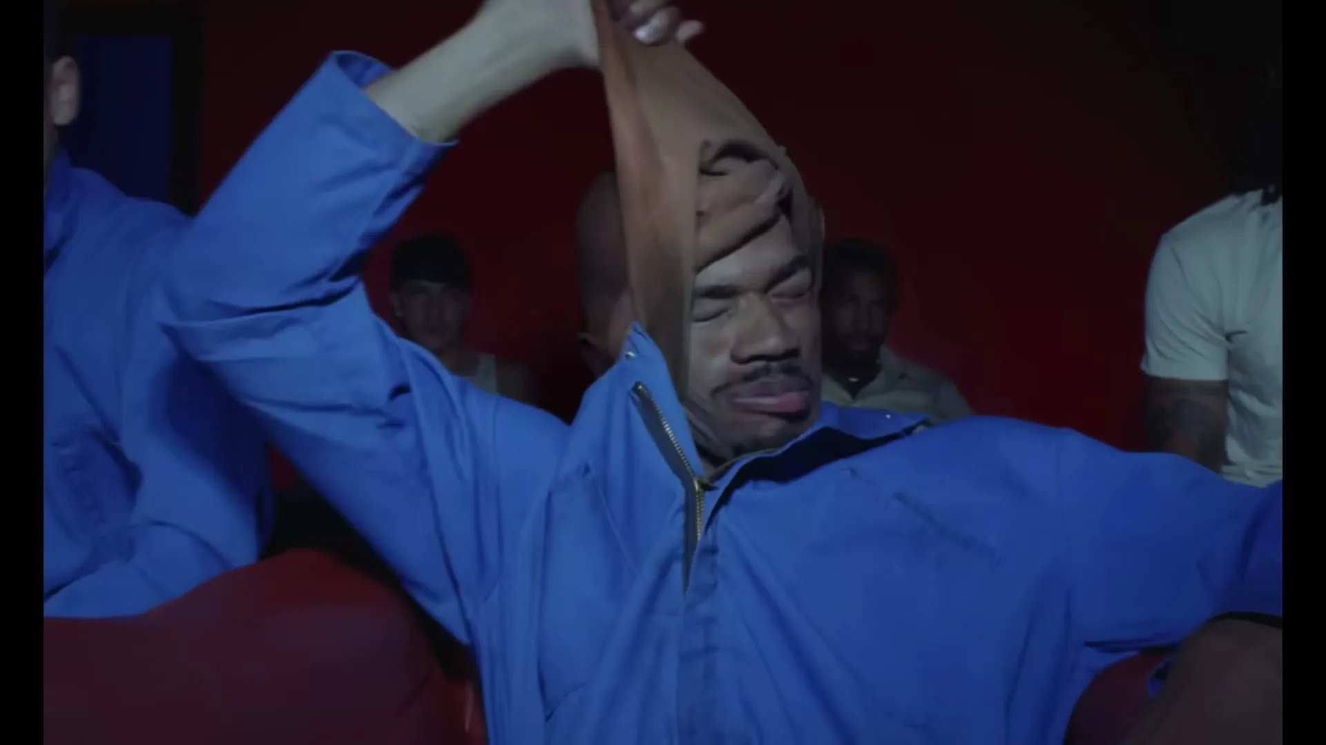 A man in a blue jumpsuit has a stocking pulled over his head, then it is removed to reveal a younger man smiling at the camera in a red room.