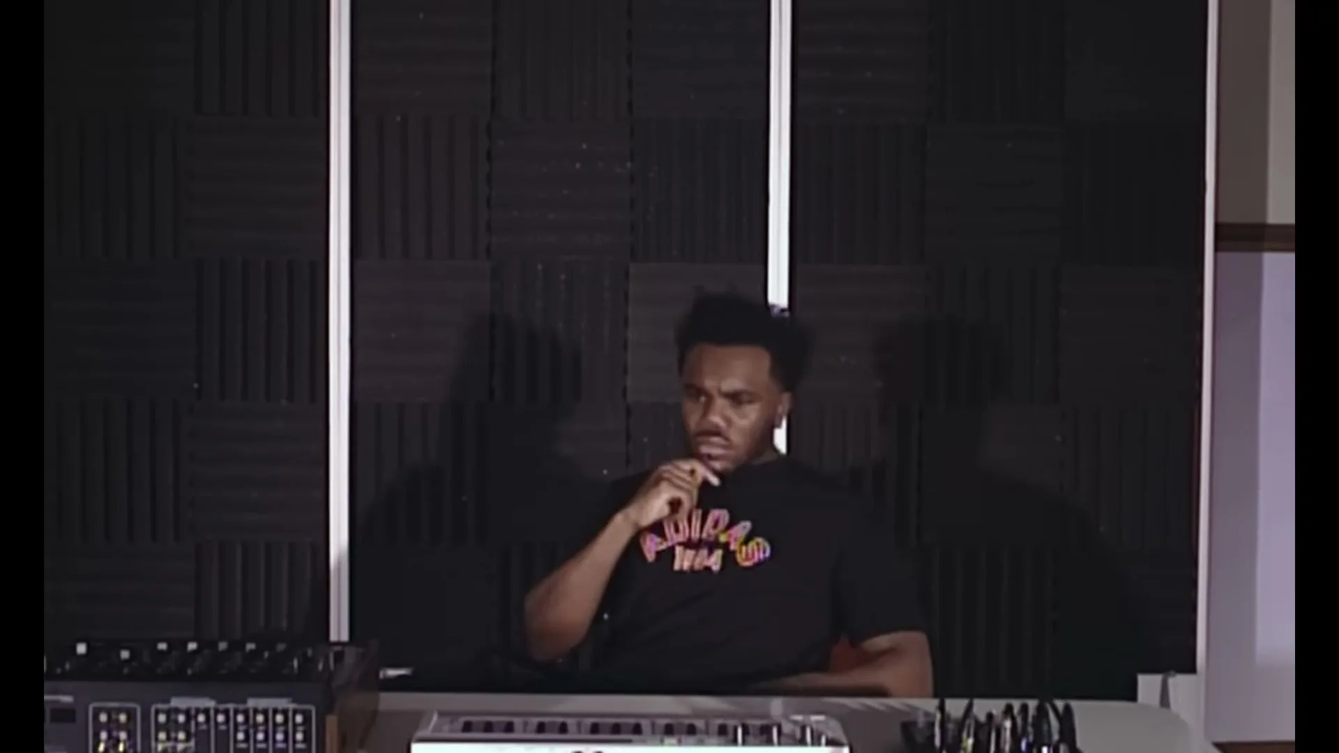 A young Black man in a black t-shirt sits at a mixing desk, looking thoughtful in a recording studio.