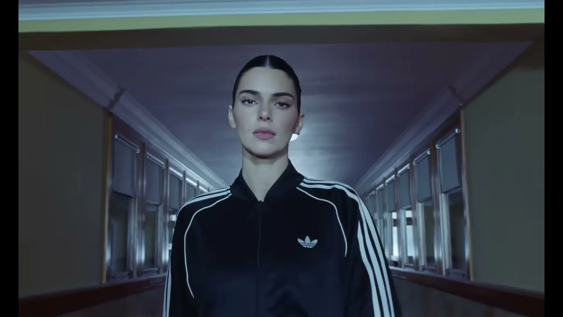 Kendall Jenner, a young woman with dark hair, wearing a black Adidas tracksuit, opens her eyes and looks directly at the camera in a dimly lit hallway.