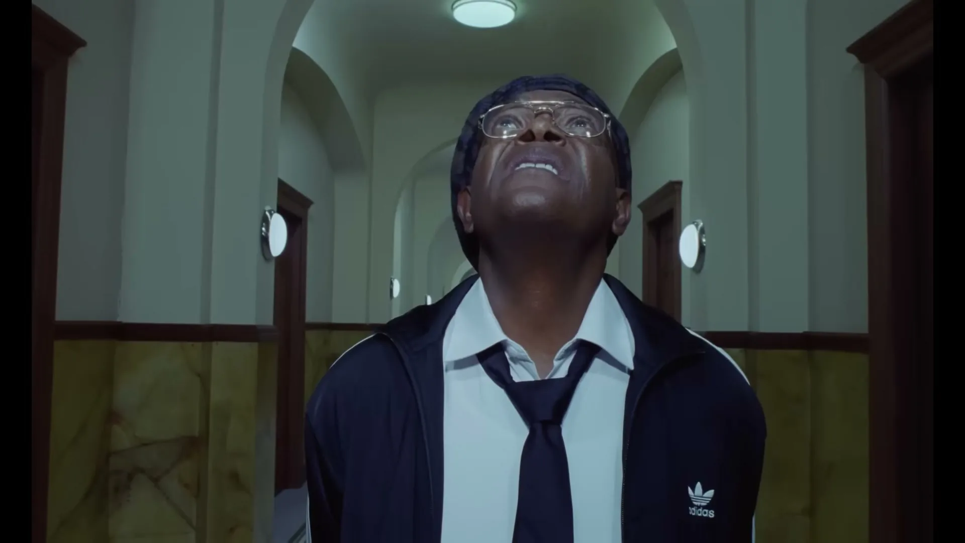 A man in a track jacket, shirt, and tie looks up with a surprised expression in a long, empty hallway.