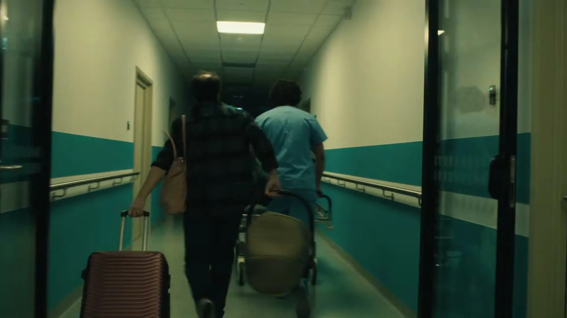 A man and a nurse rush down a hospital corridor, pushing a gurney and carrying a suitcase, as they head towards a bright light at the end of the hall.