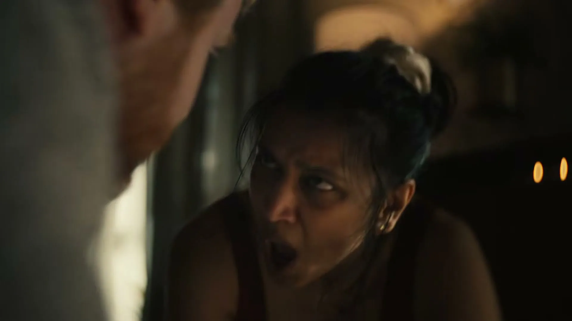 A woman in a red sports bra is seen from a medium closeup, looking up at someone off-screen with a strained expression in a dimly lit room.