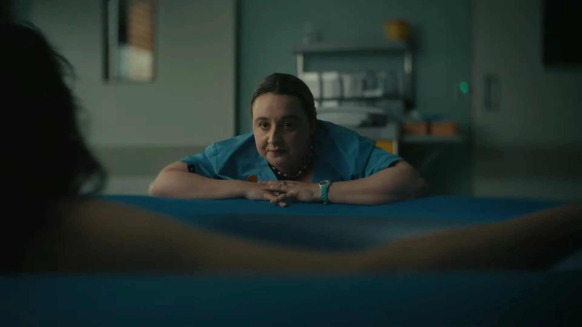 A female midwife in blue scrubs leans over a birthing pool, looking intently at a woman whose legs are visible in the foreground, in a hospital room.
