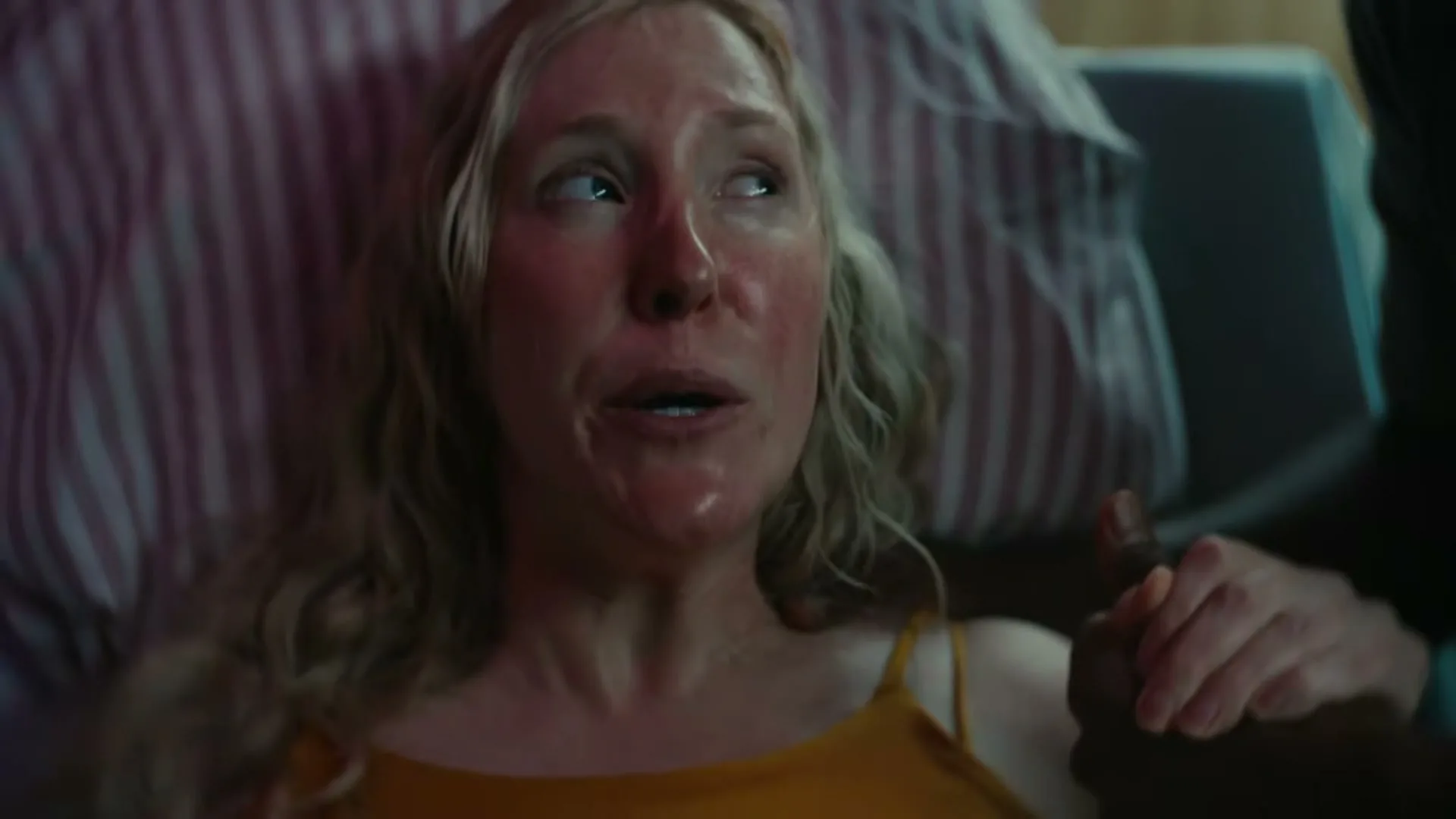 A sweaty, red-faced woman in a yellow top lies on a striped pillow, looking up and holding a man's hand in a hospital room.