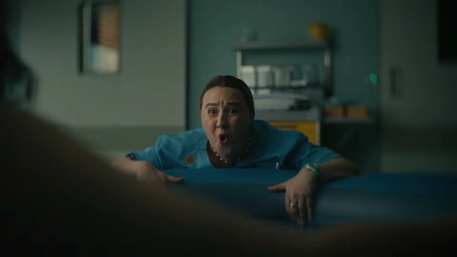 A female midwife in blue scrubs leans over a birthing pool, looking shocked and wide-eyed, in a hospital room.