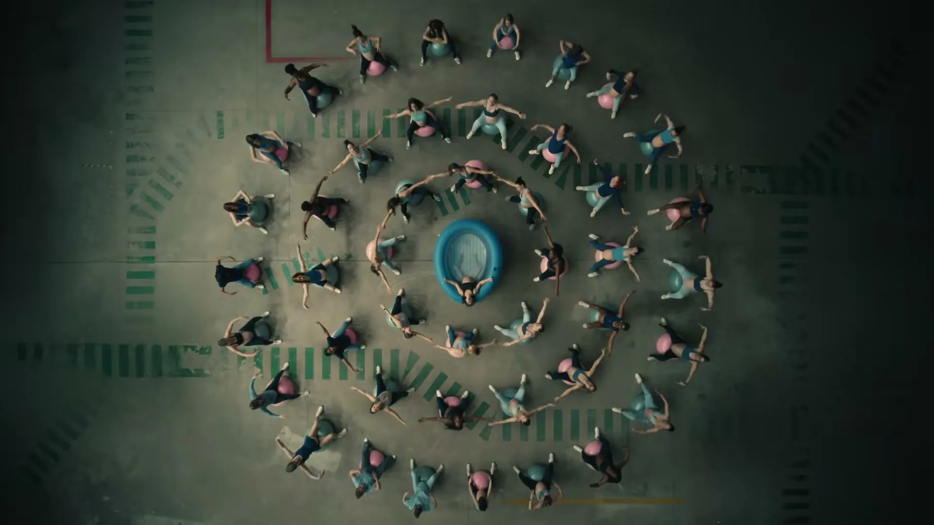 A woman in a black top lies in a blue inflatable pool, surrounded by other women on exercise balls, all pushing and grunting in a large, empty room.