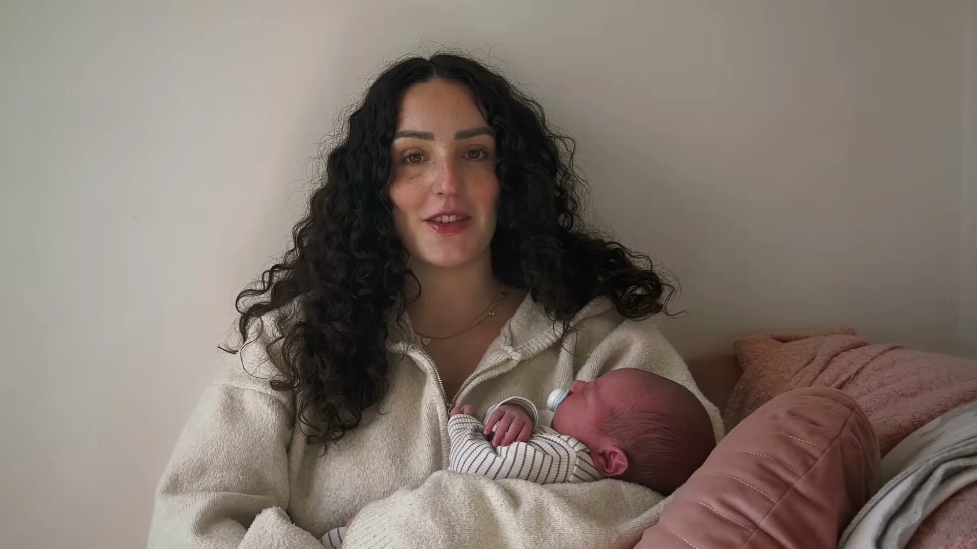 A young woman with long curly hair, wearing a cream-colored hoodie, holds a newborn baby with a pacifier in a cozy room.