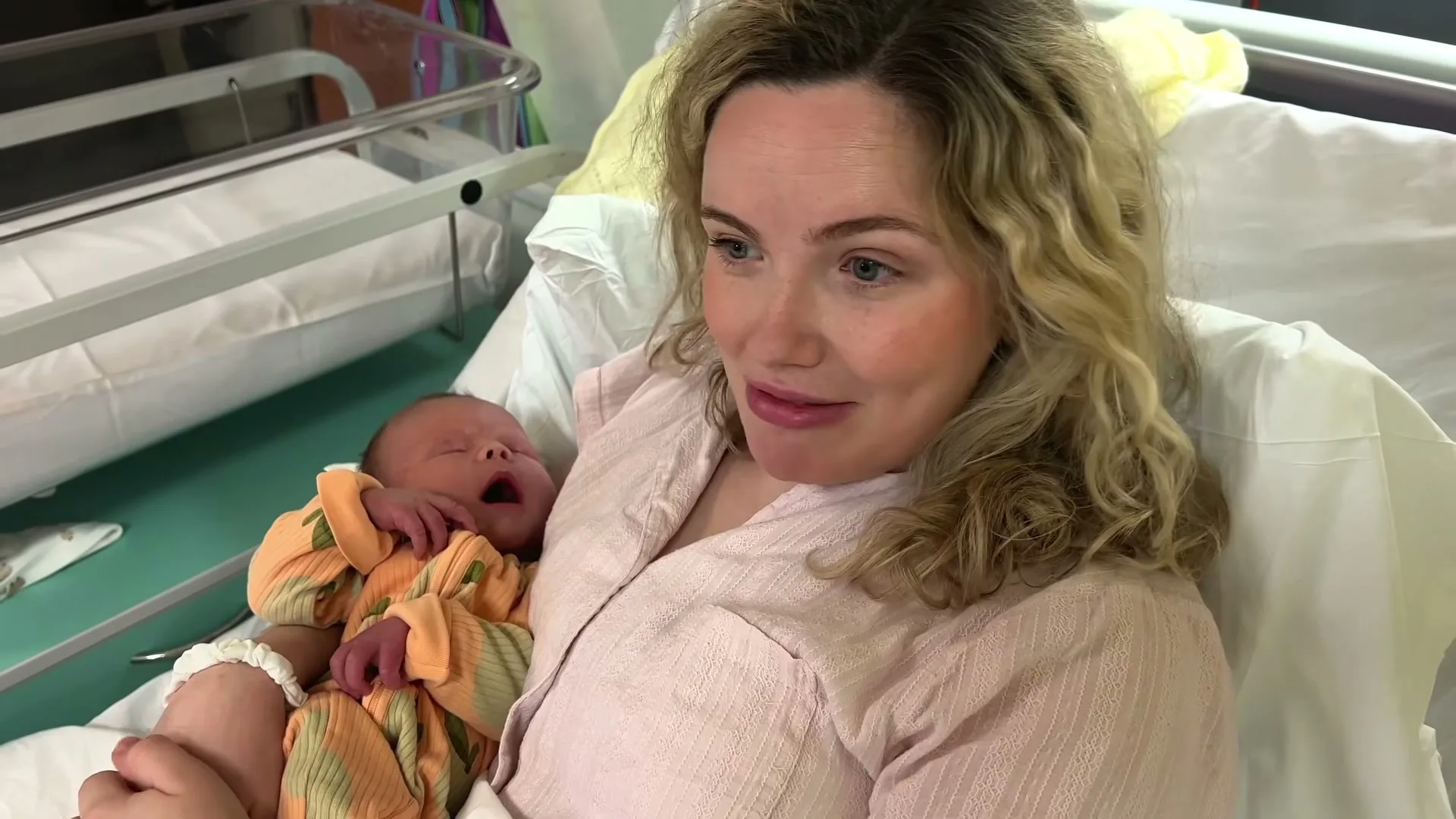 A new mother with curly blonde hair, wearing a pink robe, holds her newborn baby in a hospital bed, looking at the camera and talking.