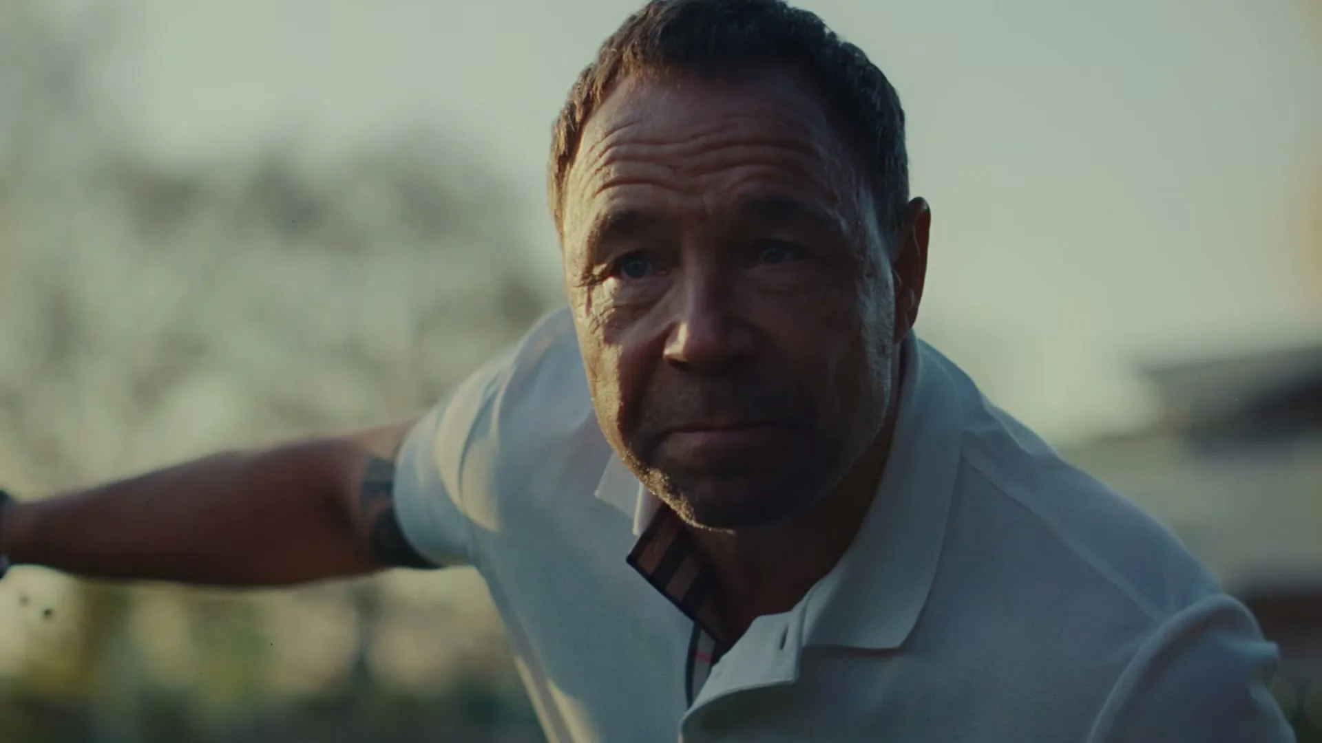 A determined man in his 40s, wearing a white polo shirt, looks intently at the camera while speaking outdoors during golden hour.