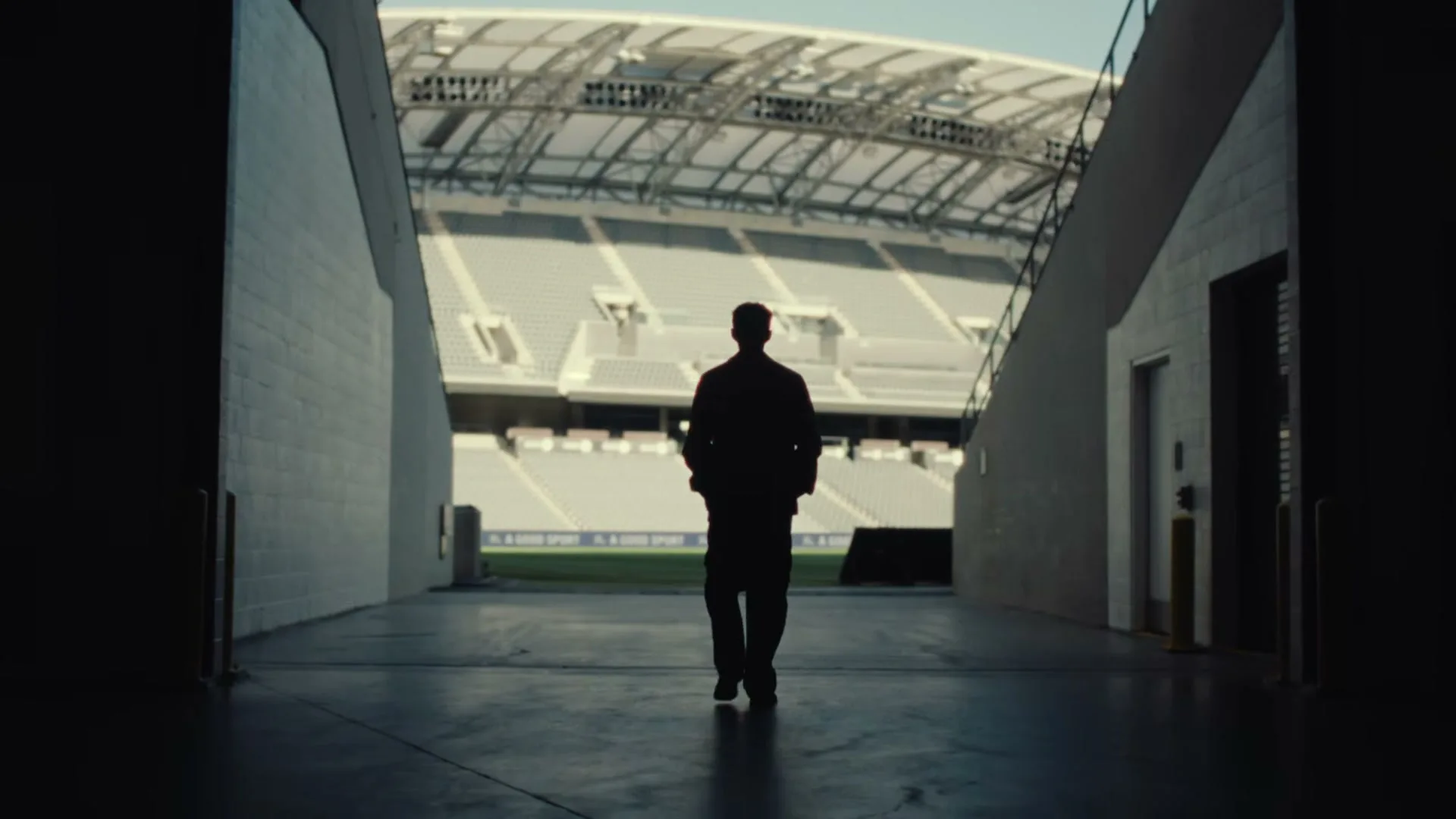 A male figure, silhouetted, walks away from the camera down a stadium tunnel towards a bright, empty stadium.