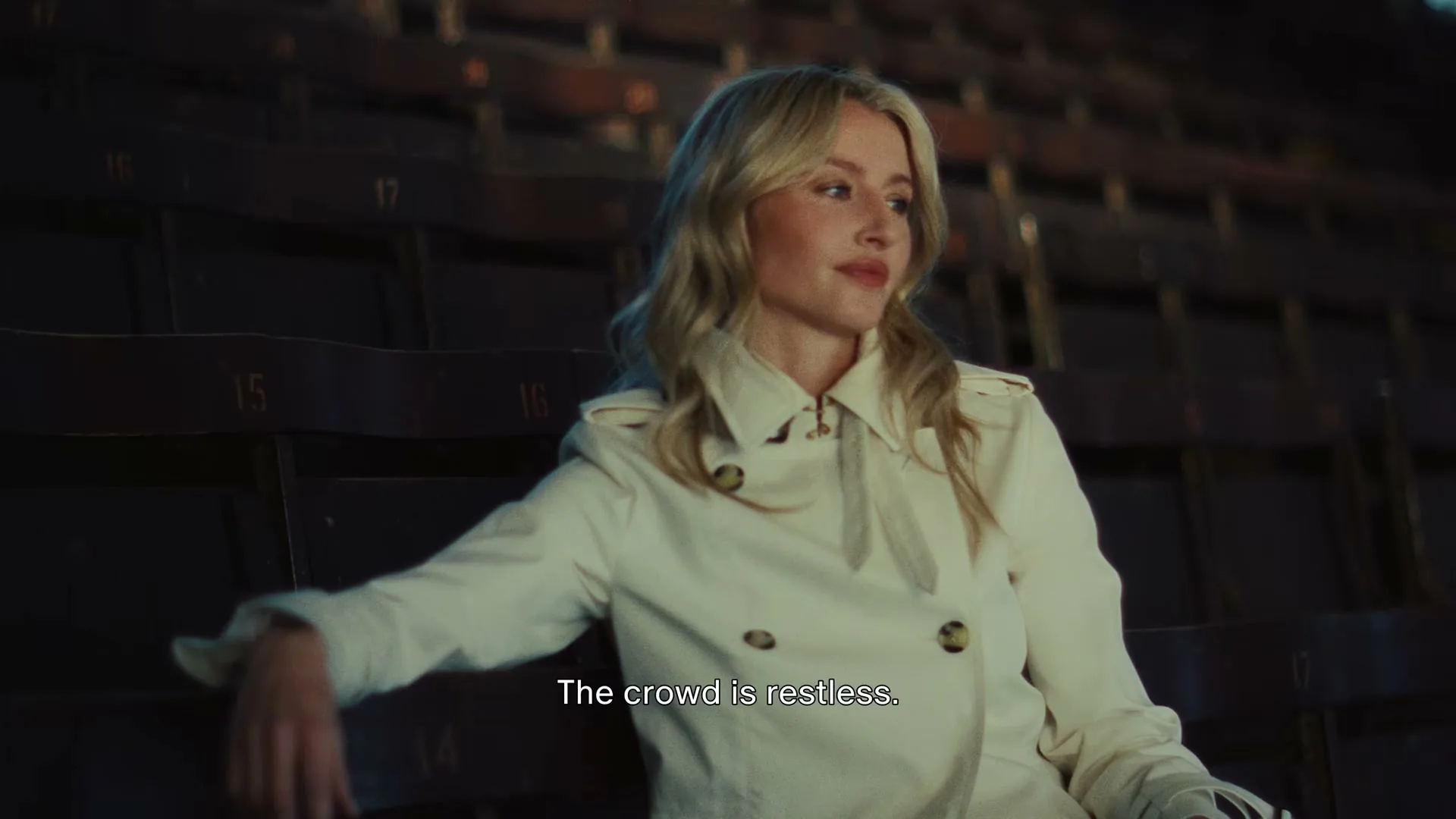 A young blonde woman in a trench coat sits alone in a dark, empty stadium, looking off to the side with a thoughtful expression.