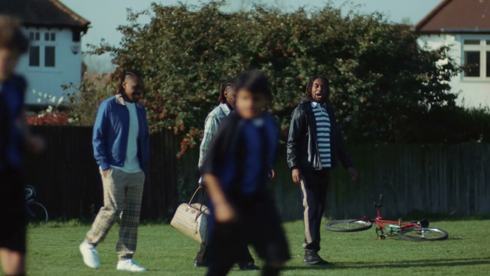 Three young men walk across a grassy field, one carrying a duffel bag, as two children in soccer uniforms run past in the foreground.