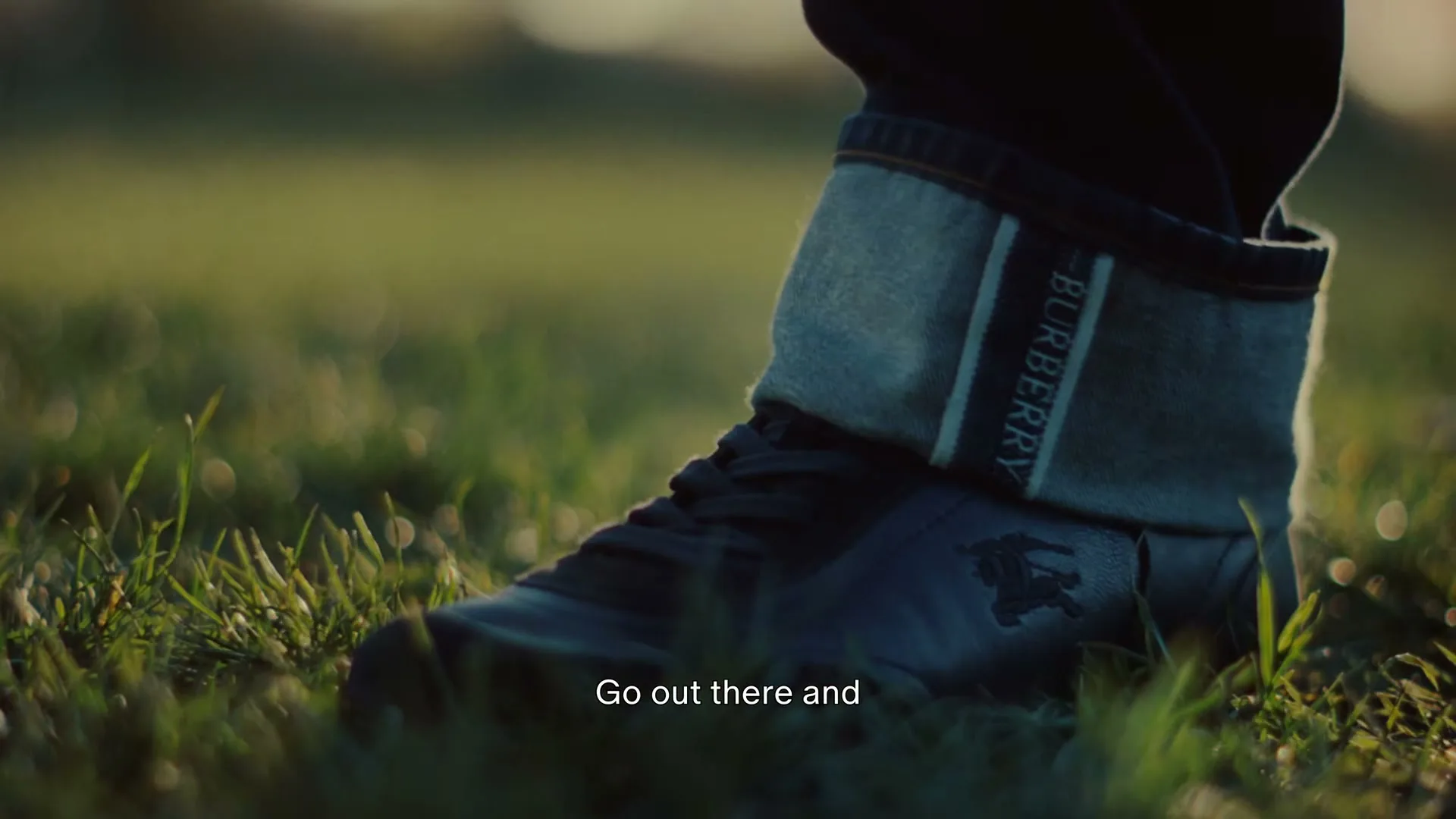A person's foot, wearing a black shoe with a Burberry logo and cuffed jeans, stands on green grass in soft sunlight.