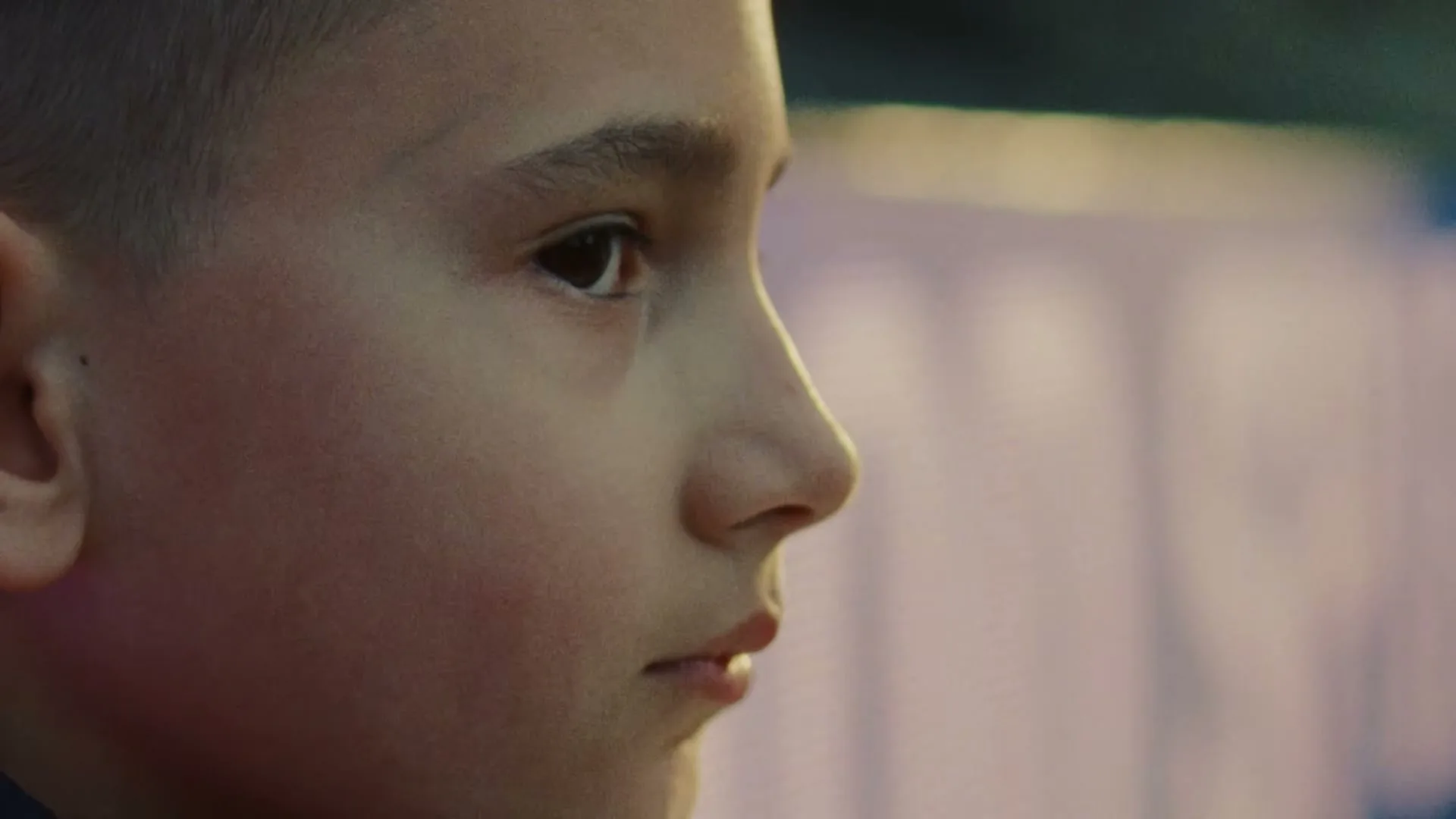 A young boy with short hair looks intently to the right, his face illuminated by soft light, in a close-up shot.