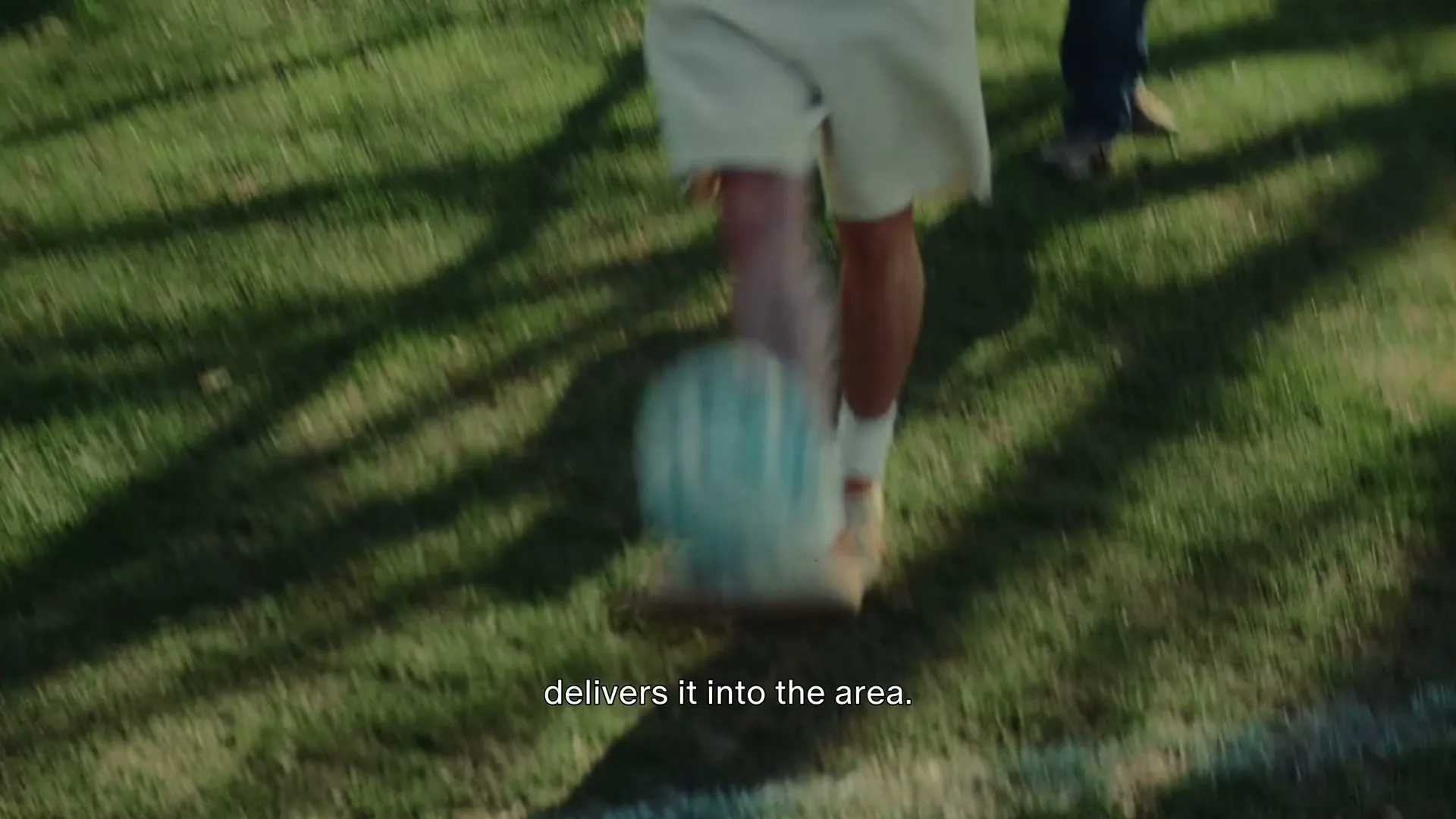 A person in light shorts and a jacket kicks a blue and white soccer ball across a grassy field, with another person's legs visible in the background.