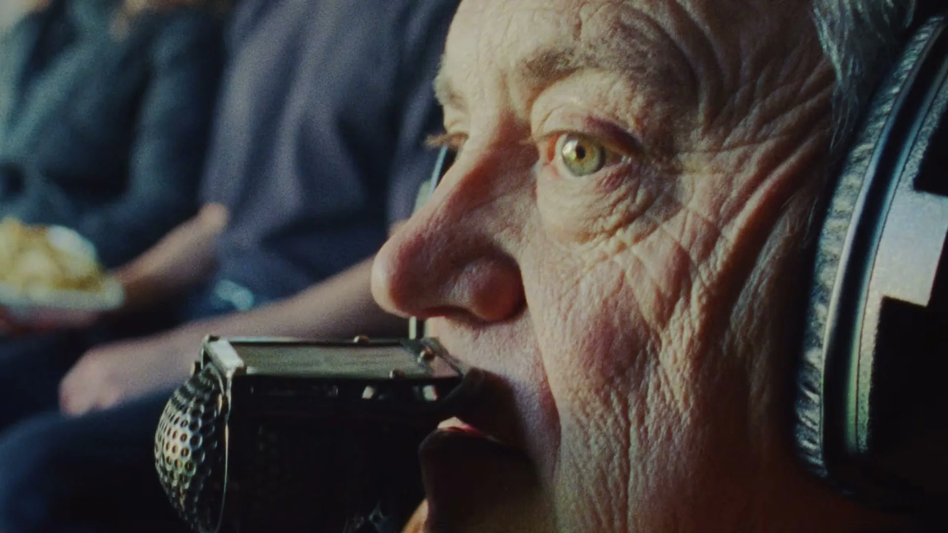 A close-up of an older person with headphones and a microphone, speaking intently, with a blurred background of other people.