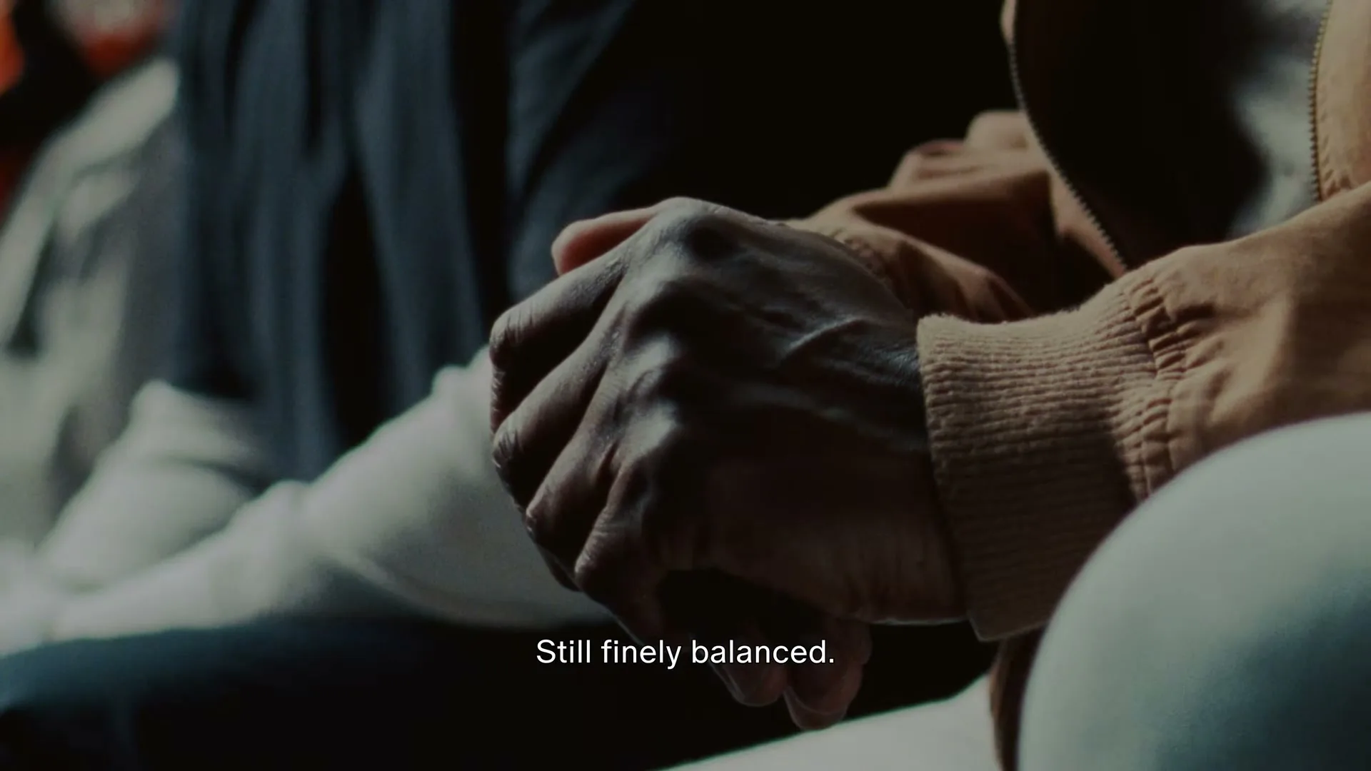 Two people's hands are clasped together, one wearing a brown jacket, in a dimly lit interior setting.