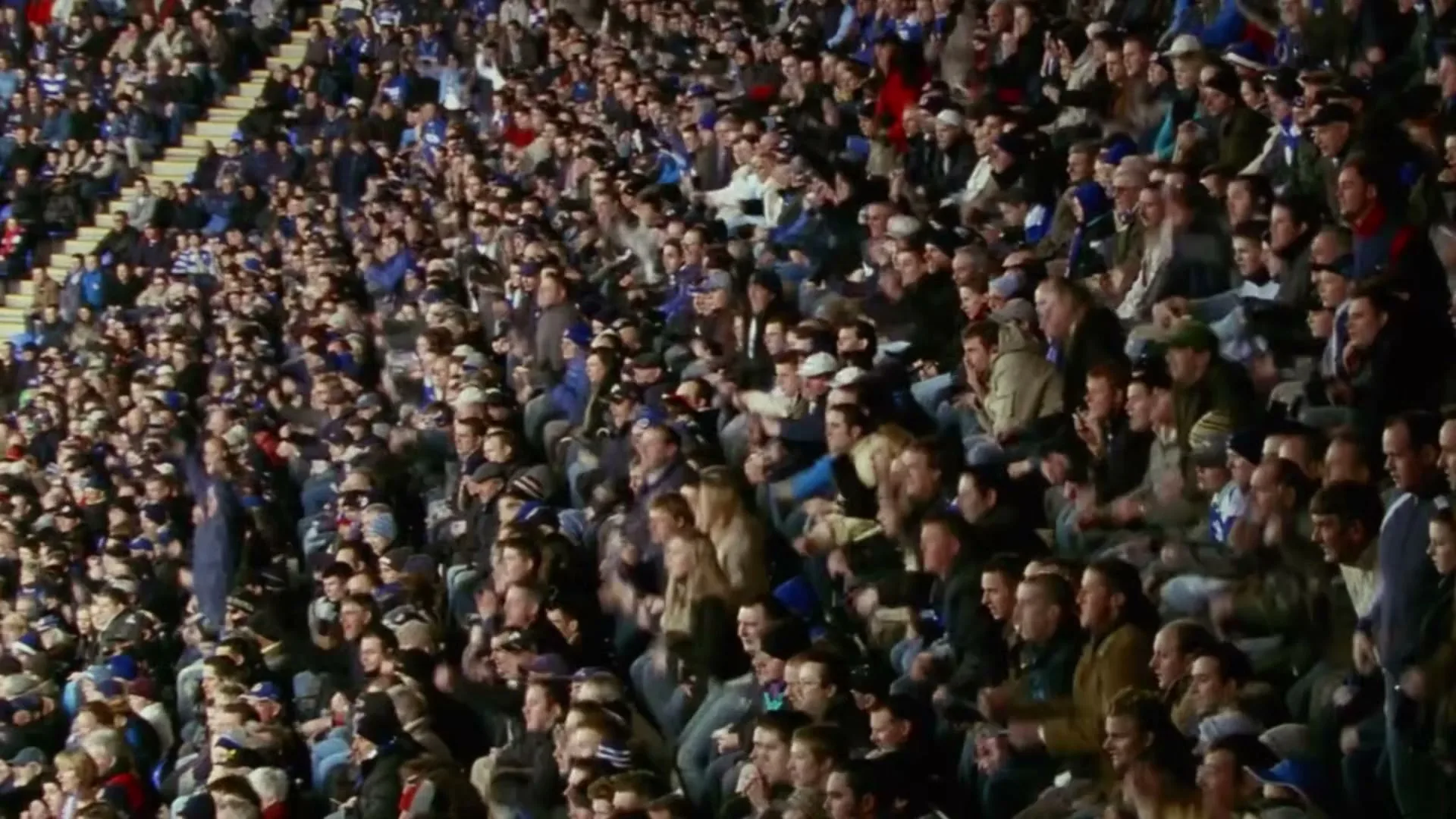 A large crowd of diverse people in casual attire cheer and raise their arms in a stadium during a sporting event.