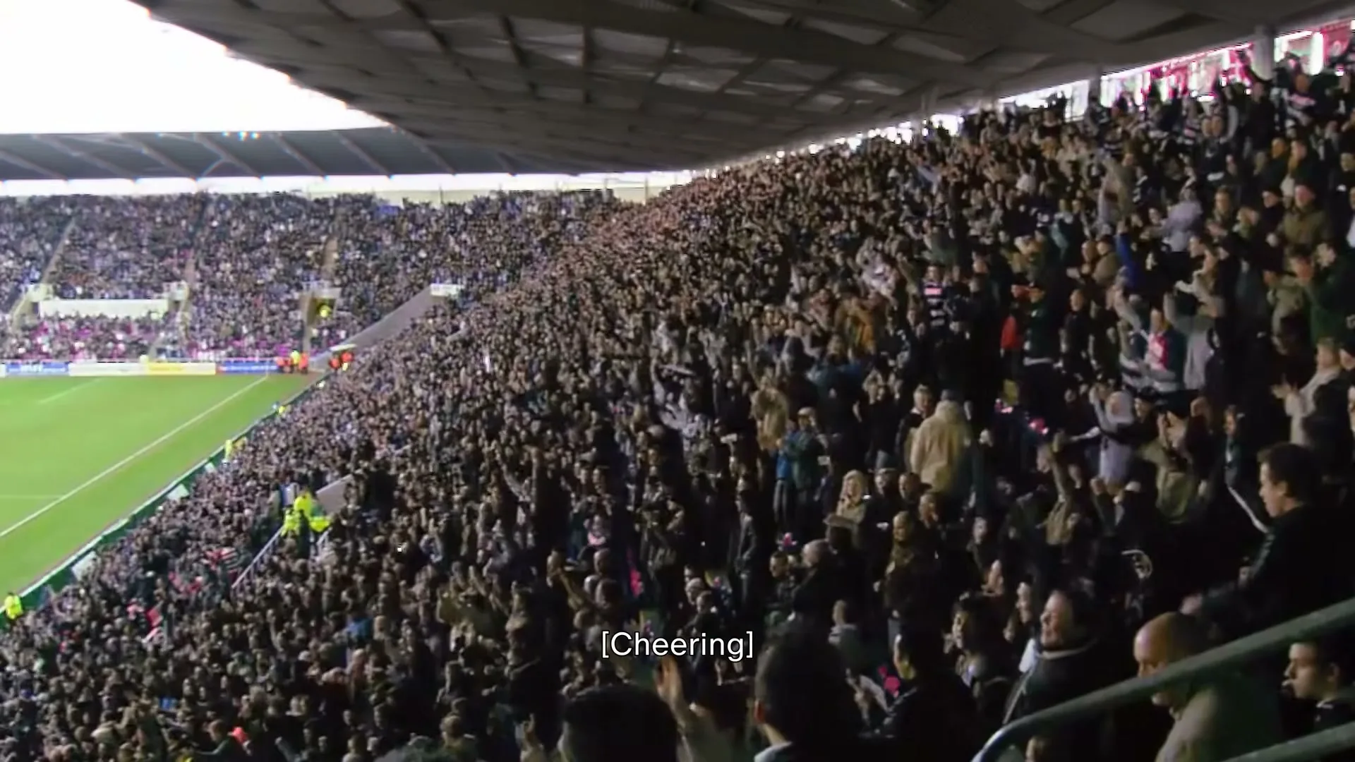 A wide shot captures a massive crowd of people cheering and clapping in a stadium, with a green field visible on the left.