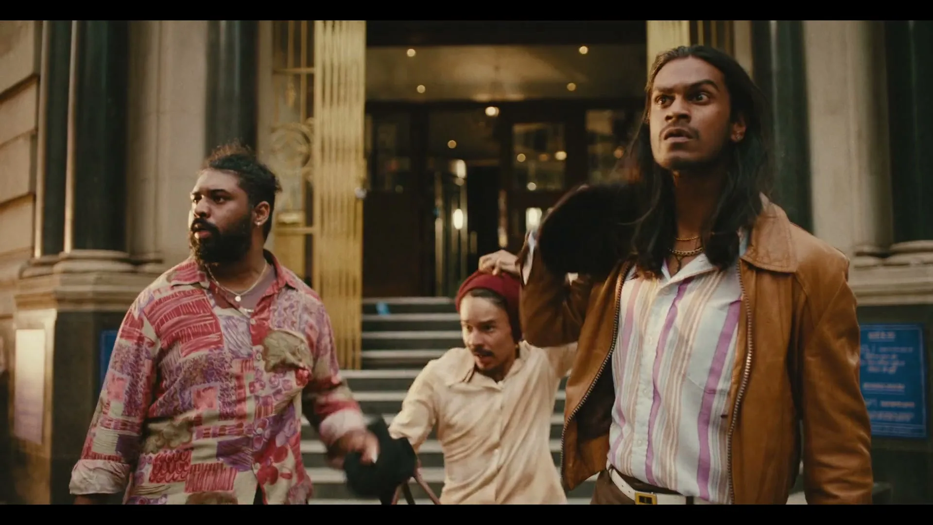 Three men, two with long hair and one with a beard, remove balaclavas as they walk away from a building entrance on a city street.