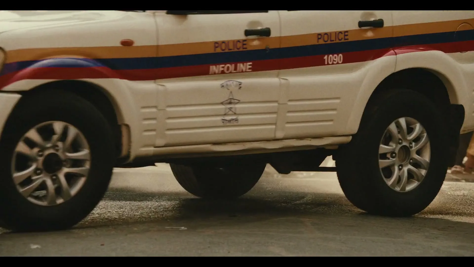 A white police SUV with red and blue stripes speeds through a city street, kicking up dust as it turns a corner past a building with "Royal Bank of India" written on it.