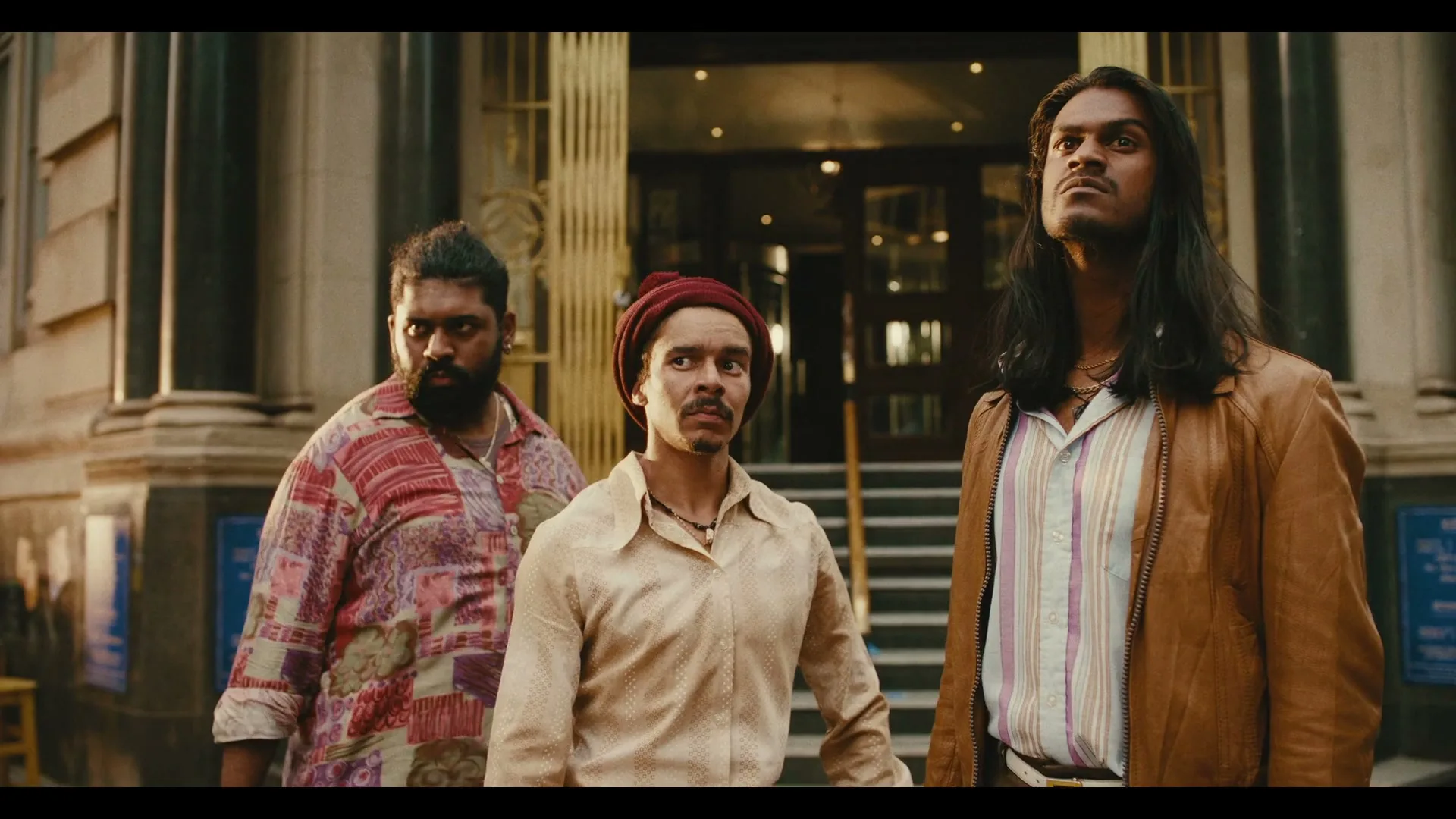 Three men, dressed in 70s-style clothing, stand on a city street, looking up with serious expressions.