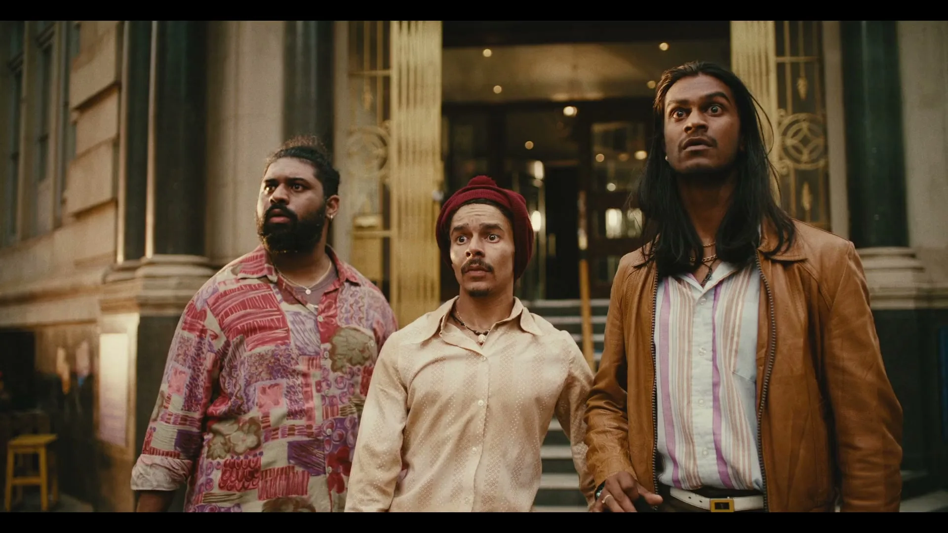 Three men, one with long hair, one with a beard, and one with a beanie, stand outside a building looking up in surprise.
