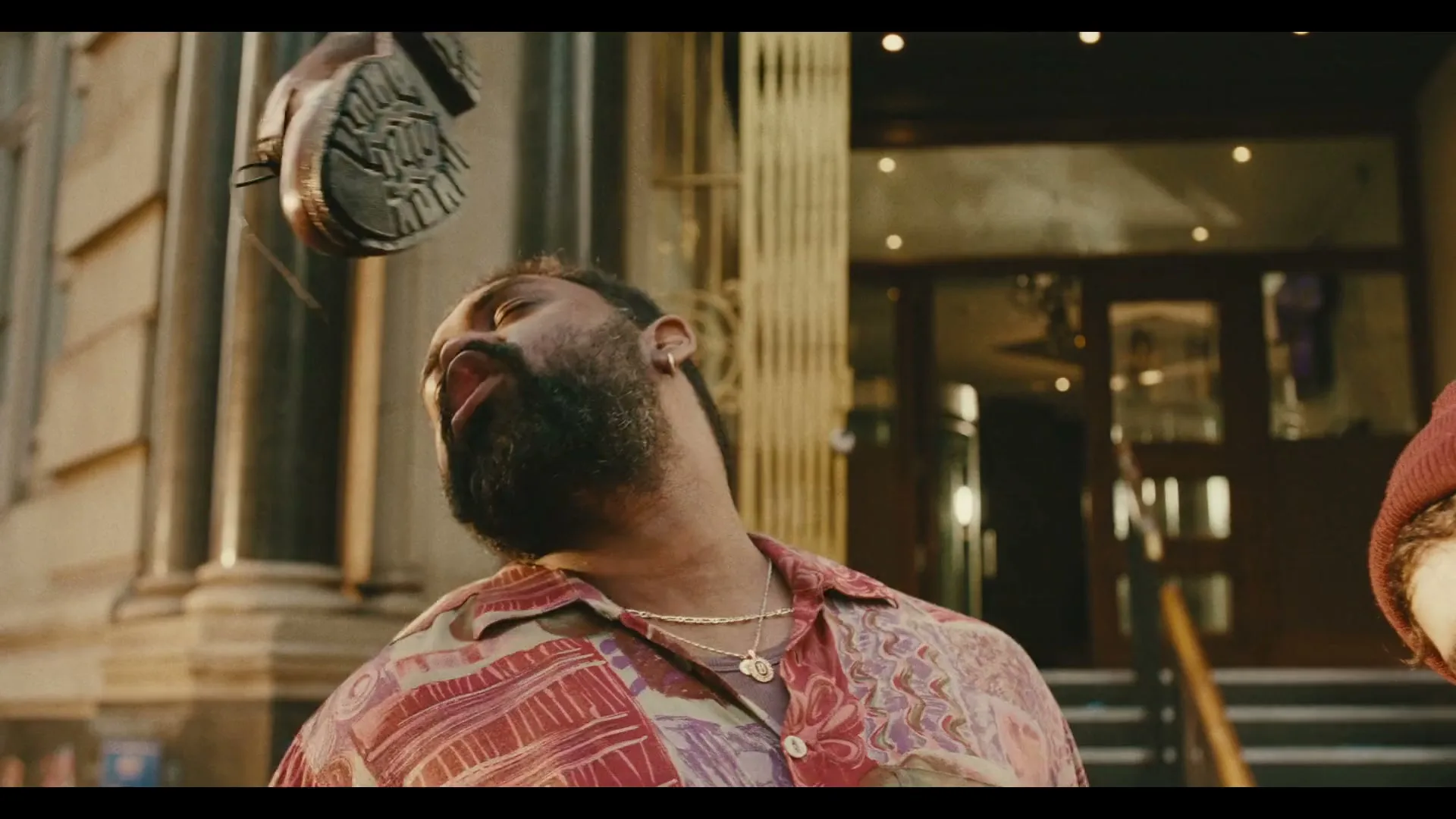A man with a beard and patterned shirt looks up in surprise as a brown shoe falls towards him on a city street.