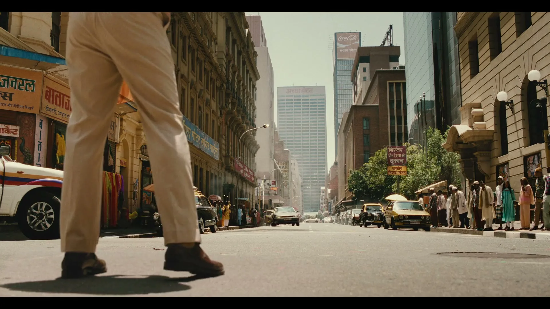 A man in tan pants and brown shoes stands in the middle of a busy city street, looking down the road at other people and cars.