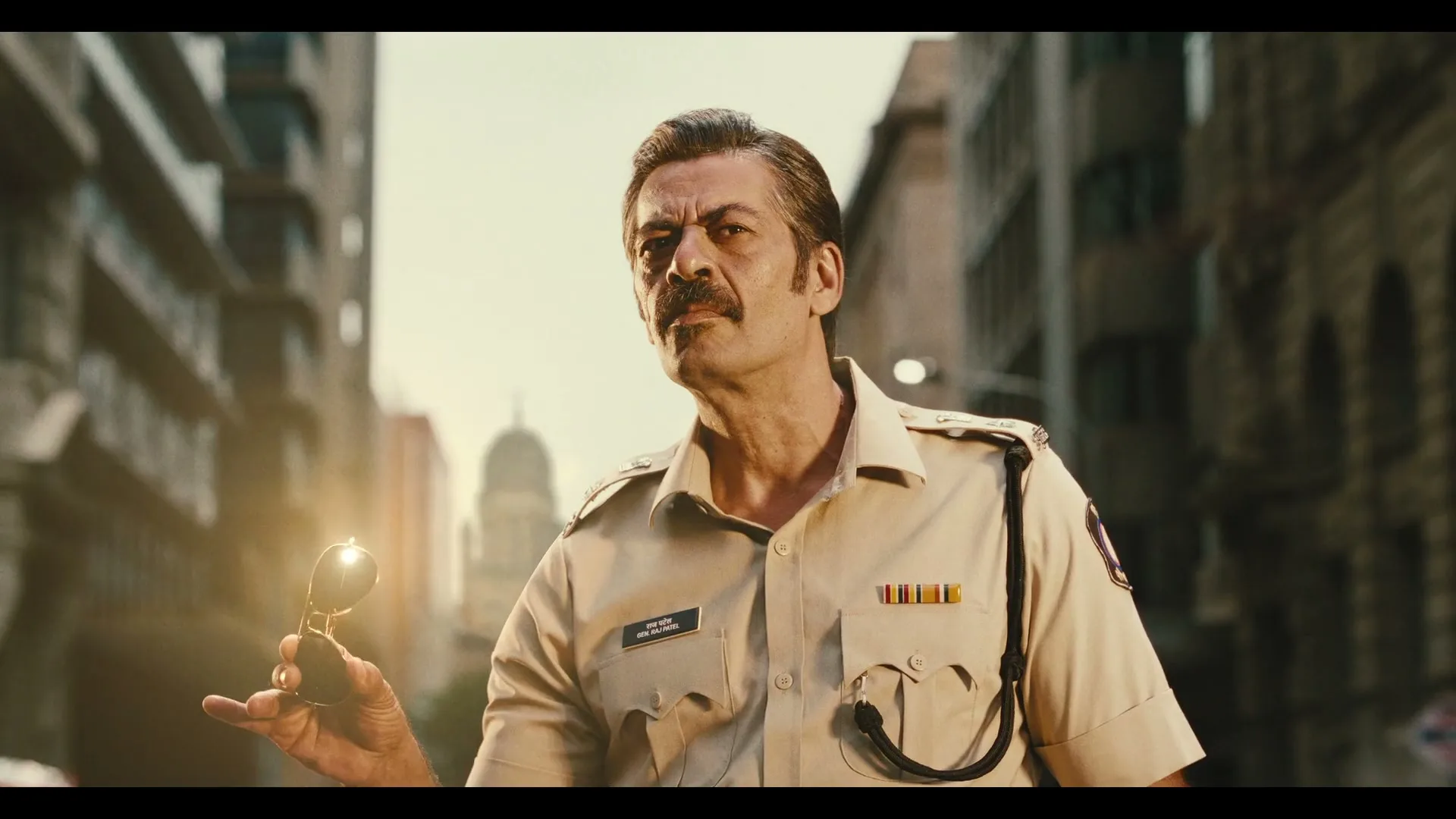 A male police officer with a mustache holds sunglasses, looking sternly at the camera on a city street.