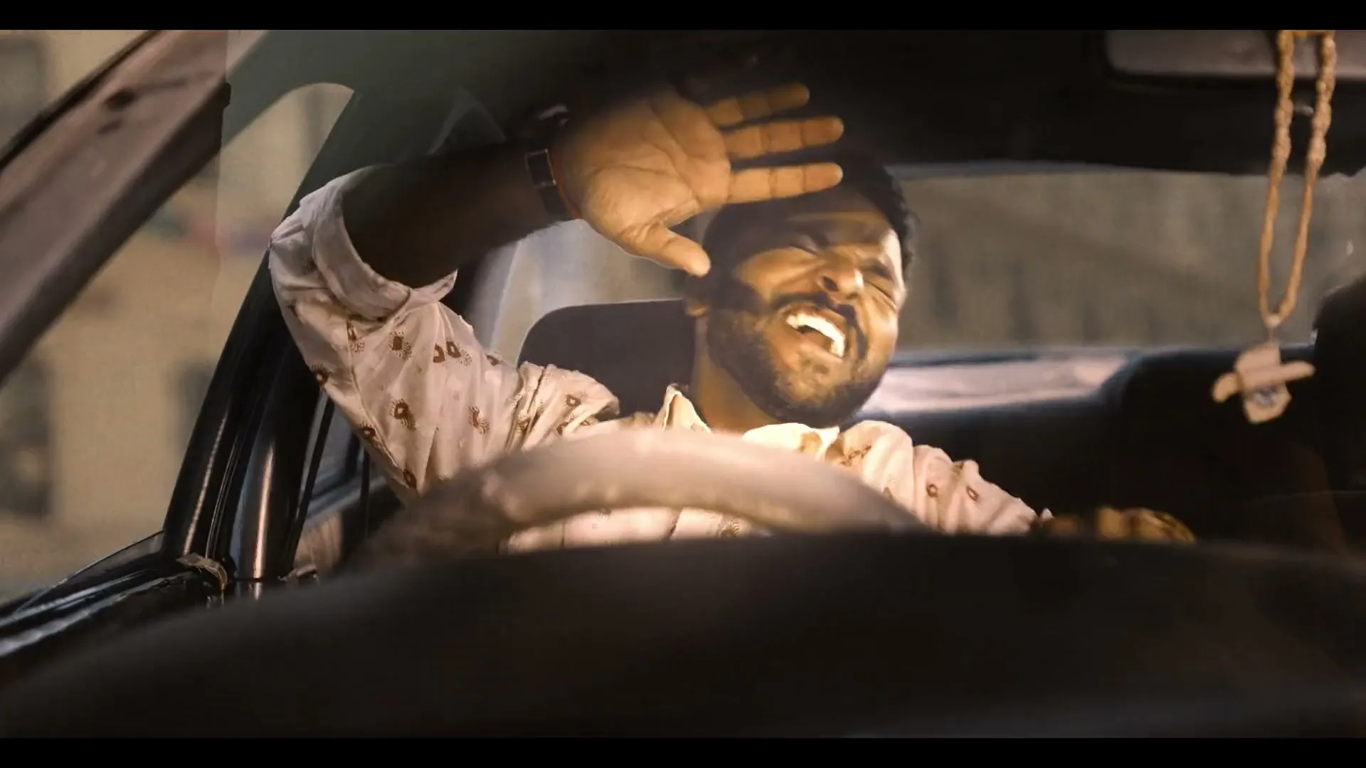 A man with a beard, wearing a patterned shirt, is driving a car and shielding his eyes from a bright light, looking distressed.