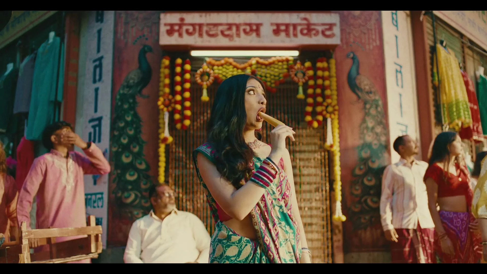 A young woman in a colorful sari eats a chicken drumstick in a bustling Indian market, surrounded by onlookers.