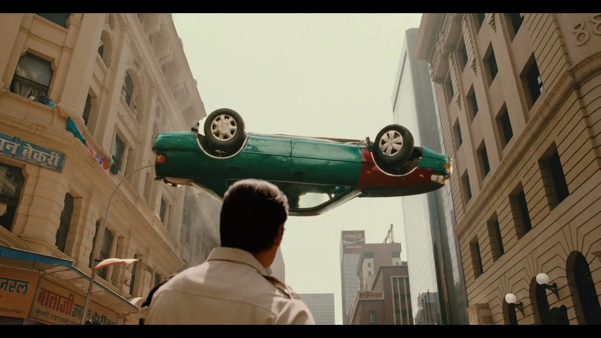 A man in a uniform looks up at a green and red car that is suspended upside down in the air between two tall buildings in a city.