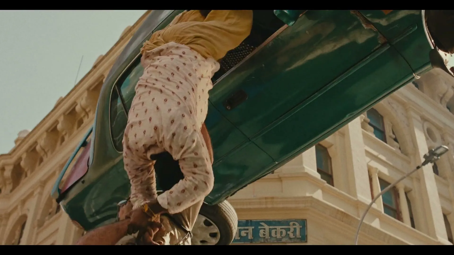 A man in a patterned shirt hangs upside down from a green car, held by another man, against a backdrop of a light-colored building.