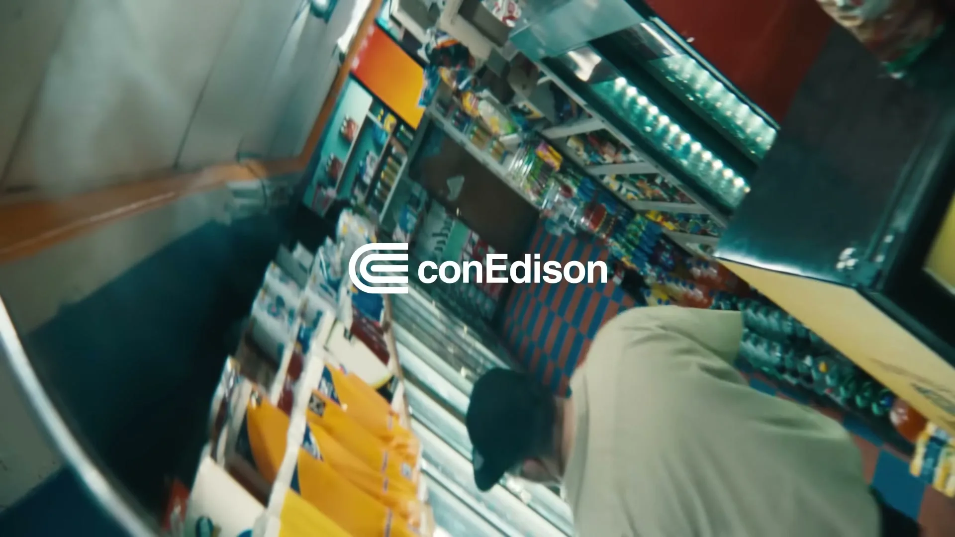 A man in a beige shirt and black cap walks through a convenience store aisle, with the Con Edison logo overlaid on the screen.