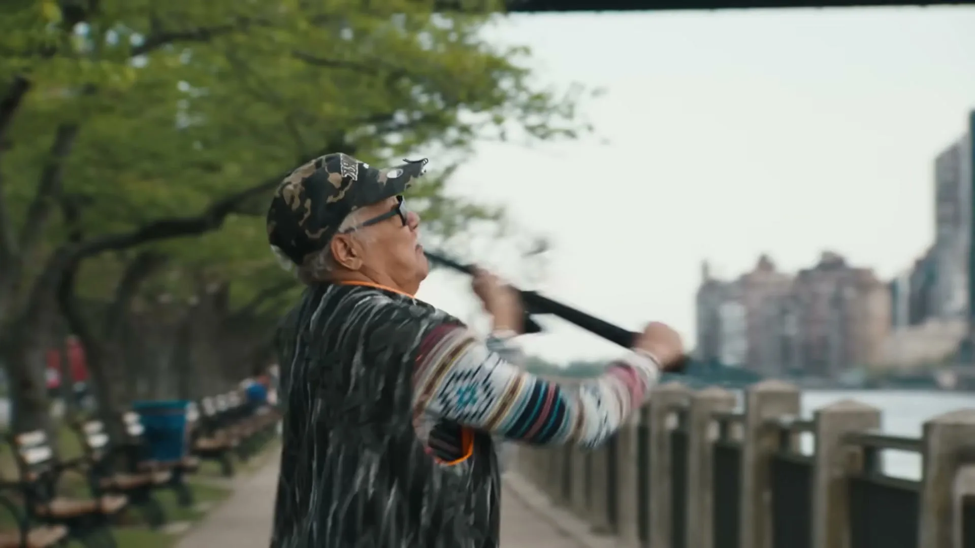 An older man in a camouflage hat and patterned sweater casts a fishing rod into a river from a park path in New York City.