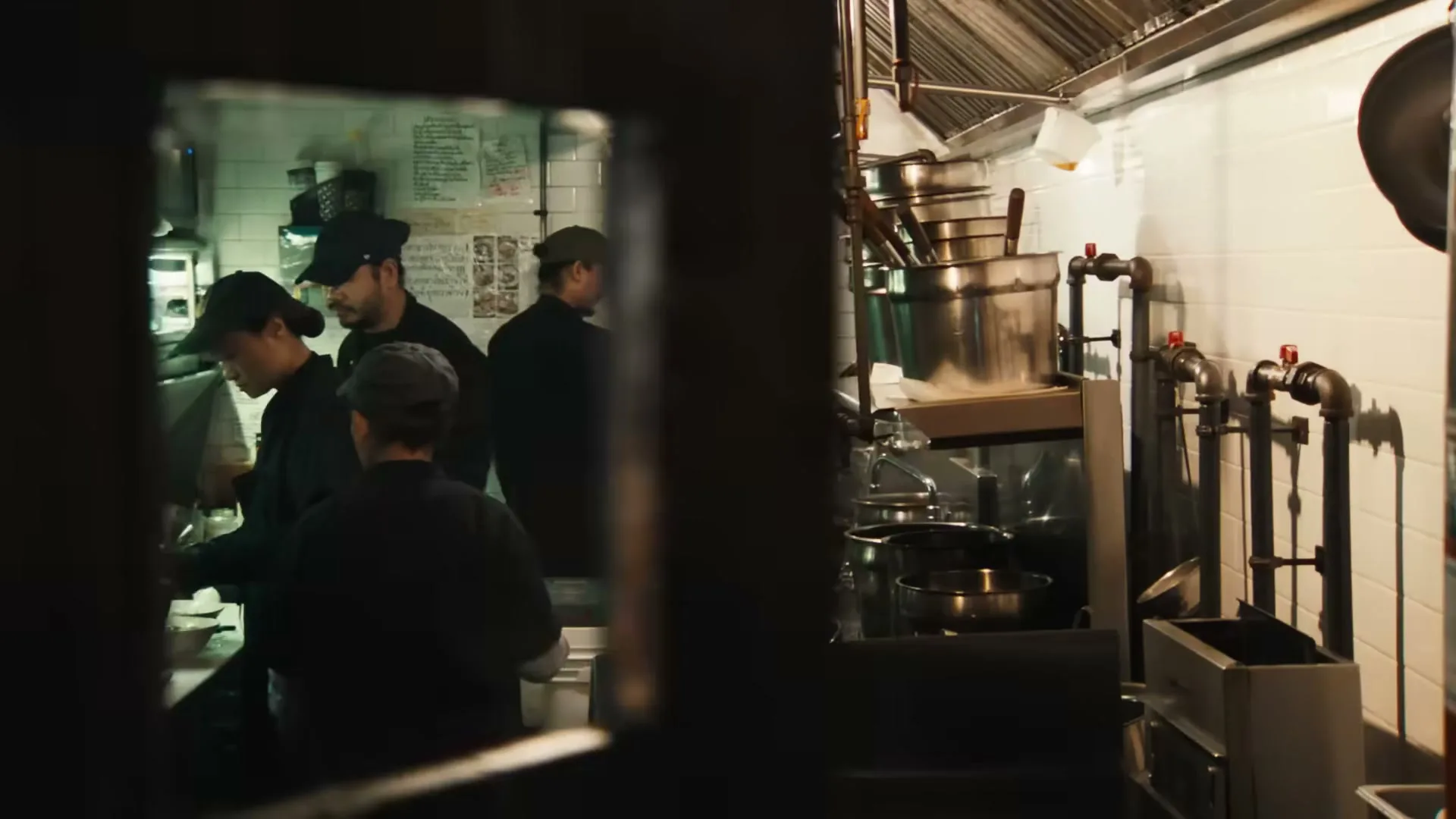 A group of chefs in black uniforms work in a busy, brightly lit commercial kitchen, seen through a doorway.