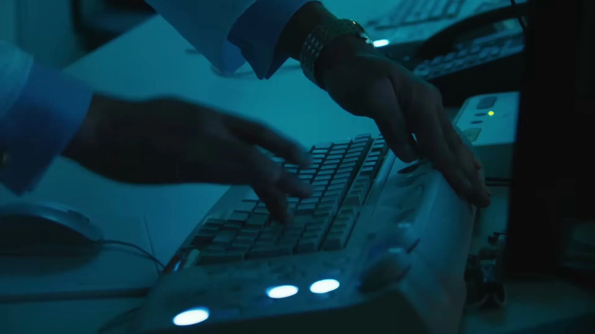 A person's hands, wearing a white shirt and a watch, type on a computer keyboard in a dimly lit room.