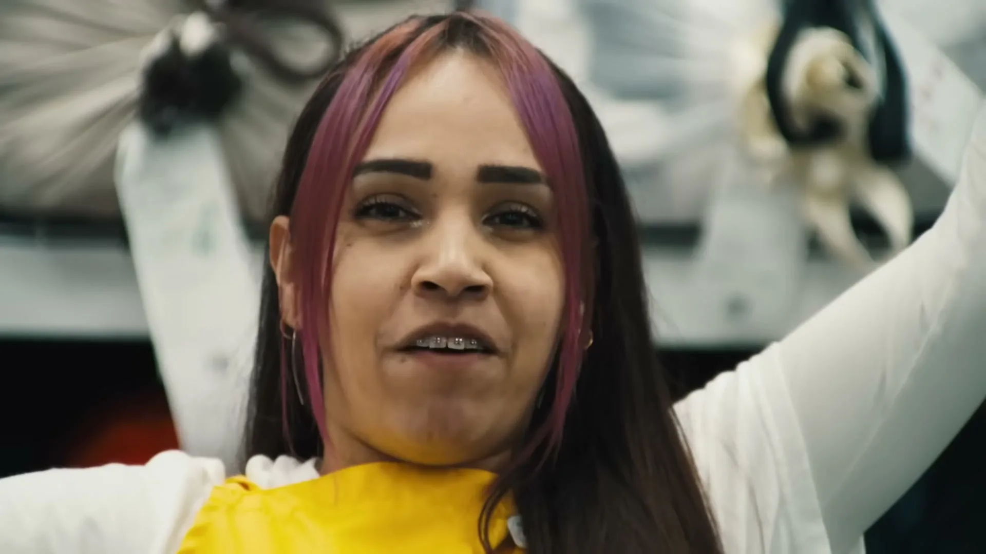 A young woman with pink-streaked hair and braces smiles at the camera, wearing a yellow top and white sleeves, in an interior setting.