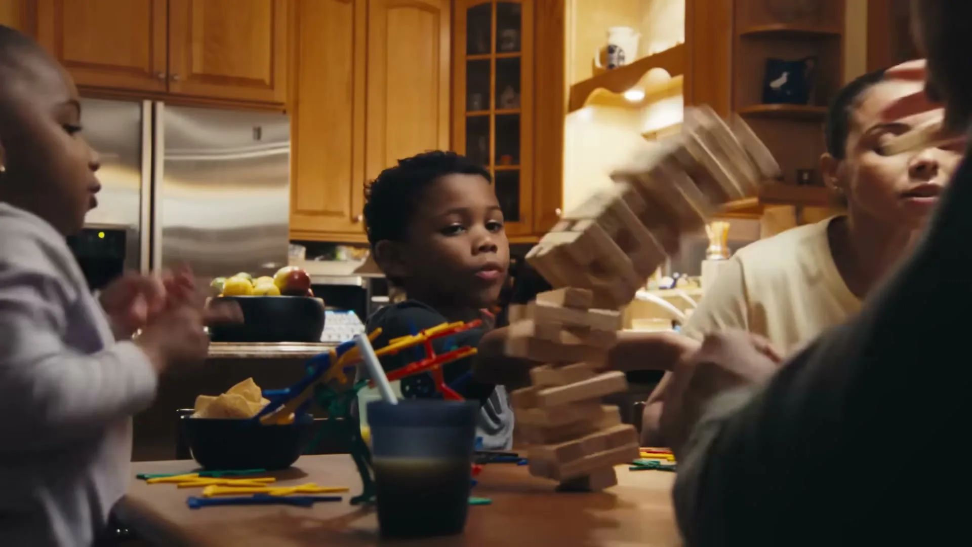 A young Black boy plays Jenga with his family at a kitchen table, and the tower collapses.