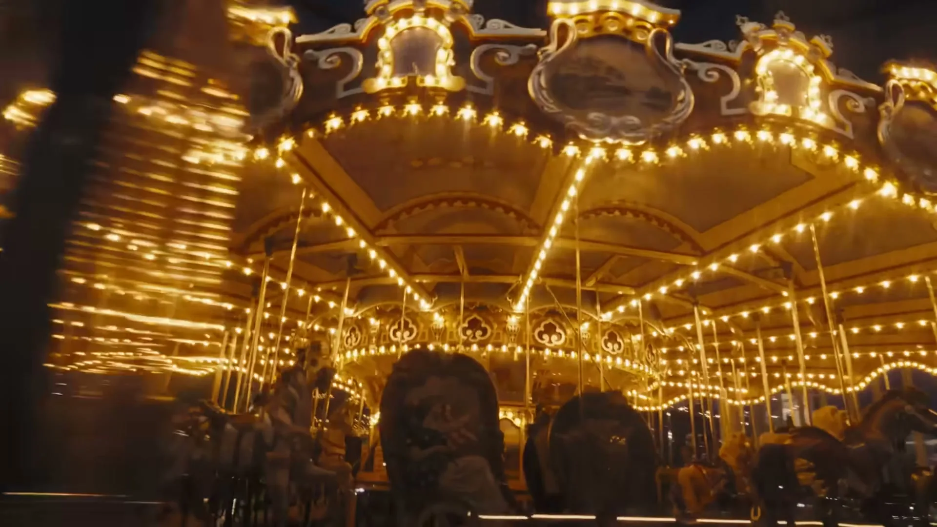 A brightly lit carousel spins at night, with blurred figures of people visible among the horses, creating a dynamic and festive scene.