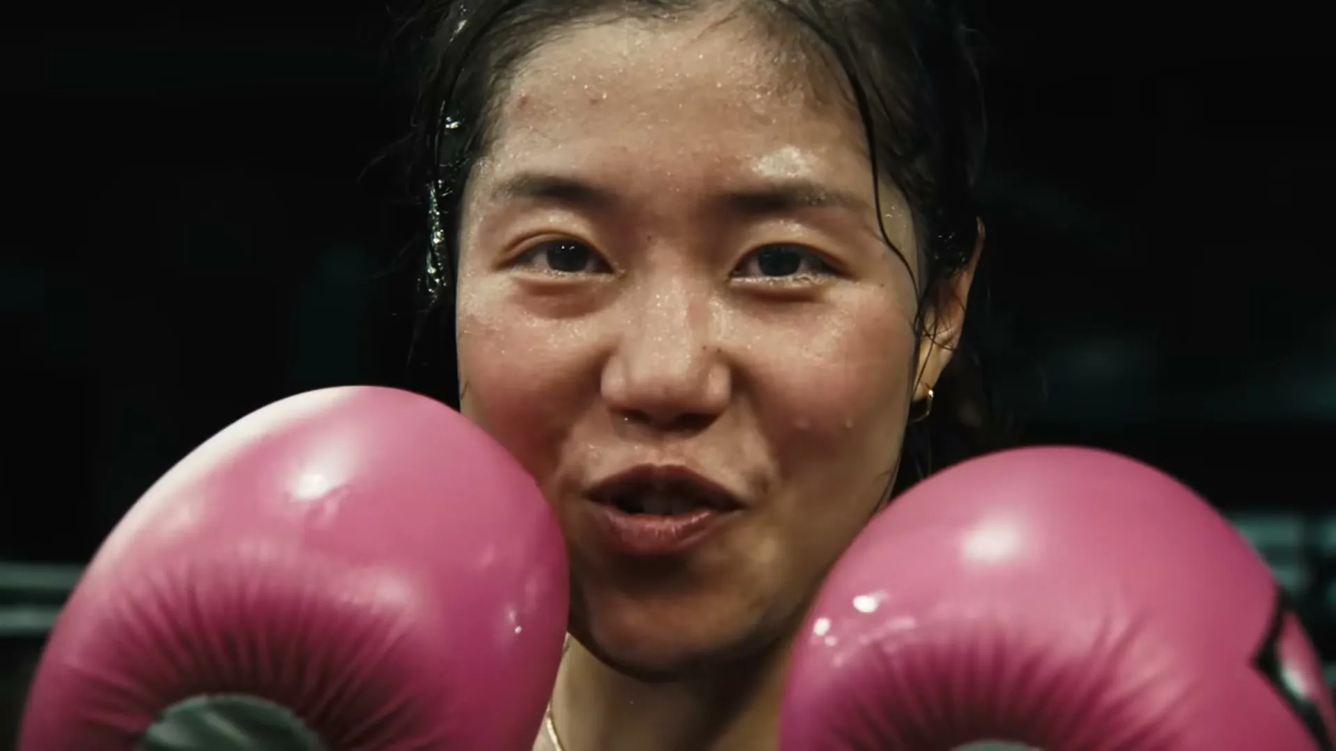 A determined female boxer, sweating and wearing pink gloves, looks directly at the camera in a boxing ring.