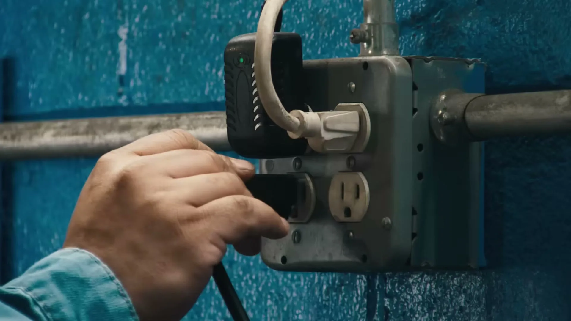 A person's hand plugs a black cord into a wall outlet on a blue wall in an interior setting.