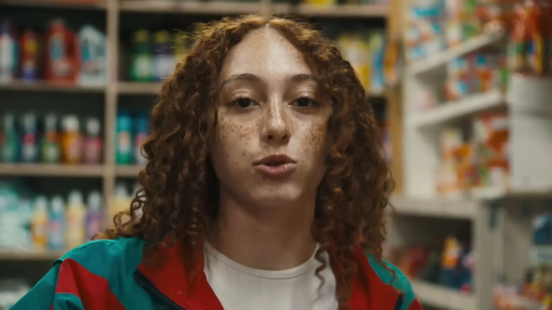 A young woman with curly red hair and freckles speaks directly to the camera in a grocery store aisle.