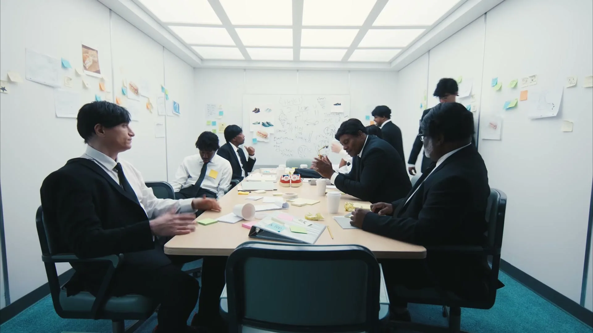 A group of men in black suits and white shirts sit around a conference table in a sterile white room, some looking bored or tired, while one man stands and addresses them.