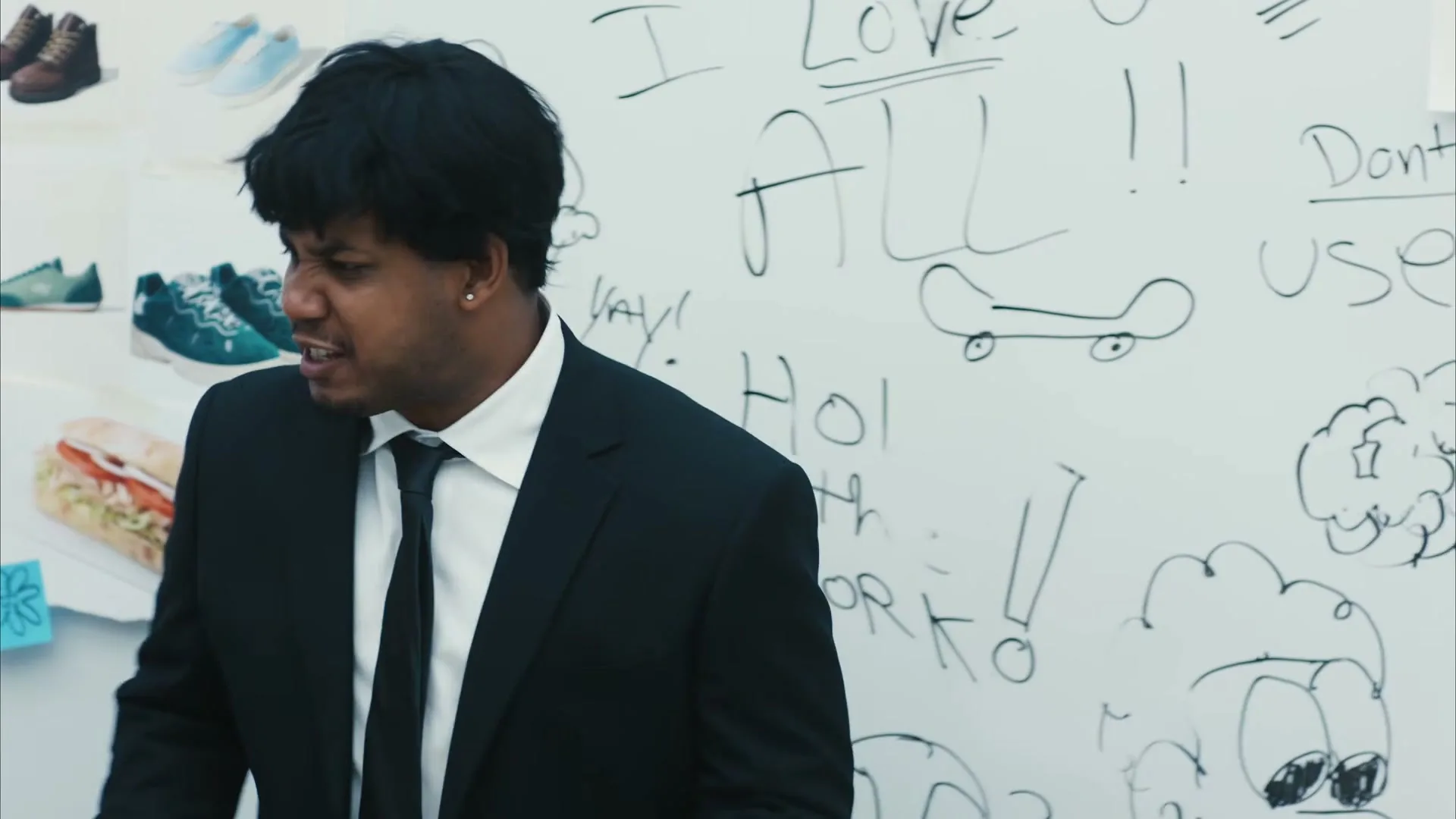 A man in a black suit and tie stands in front of a whiteboard with drawings and text, looking frustrated.