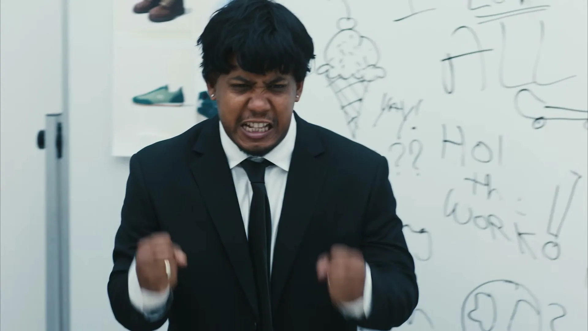 A man with dark hair and a black suit yells angrily, clenching his fists in front of a whiteboard with drawings and text.
