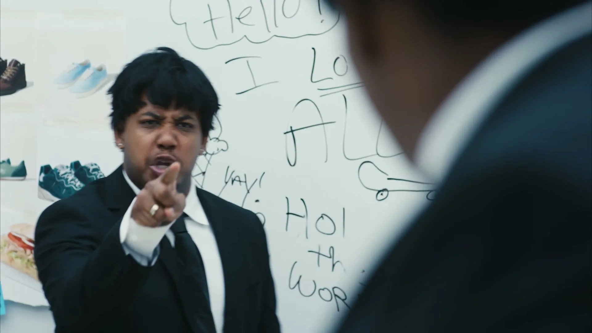 A man in a black suit and tie, with dark hair, stands in front of a whiteboard with drawings, pointing and looking angry at another person.